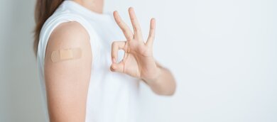 TUEV_Rheinland_Pressemitteilung_Grippeimpfung.jpg Das Bild zeigt einen Arm mit einem Pflaster, das auf eine kürzlich erfolgte Grippeimpfung hinweist. Die Hand daneben formt ein "Okay"-Zeichen, was eine positive Botschaft vermittelt.