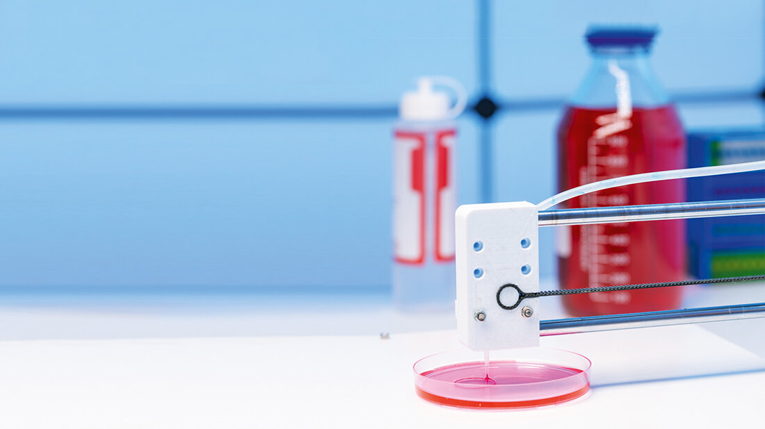 3D-Bioprinting Ein 3D-Druckkopf gibt in einem Labor eine rosafarbene Flüssigkeit in eine Petrischale ab, während im Hintergrund unscharf Flaschen und Gefäße mit Nährlösungen zu sehen sind.