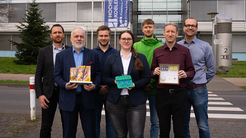 forschungsgruppe_wear-a-lab.jpg Eine Gruppe von 7 Personen einer Forschungsgruppe vor einem Gebäude mit Modellen in den Händen, Fahne der Hochschule Koblenz im Hintergrund