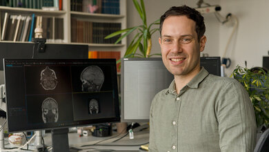 NiclasHagen_MRT_Mukoviszidose.jpg Mann in Büro am Schreibtisch, auf einem Bildschirm hinter ihm sind MRT-Aufnahmen zu sehen, Blumen und Bücherregale im Hintergrund.