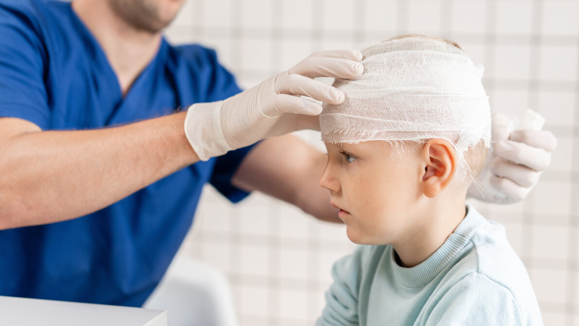 AdobeStock_279699282.jpeg Das Bild zeigt eine medizinische Situation, in der ein Kind von einem Fachmann behandelt wird. Der Arzt trägt Handschuhe und legt einen Verband um den Kopf des Kindes, was auf eine Verletzung oder eine postoperative Versorgung hinweist.