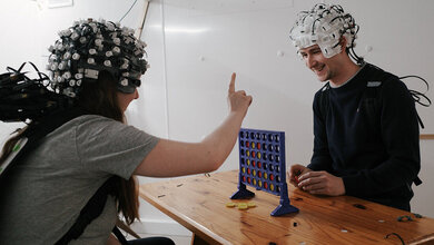 Studie_Neuroimaging.jpg Probanden einer medizinischen Studie beim Vier-gewinnt-Spiel ab einem Holztisch mit Messsonden am Kopf.