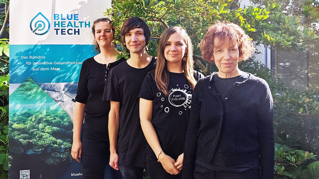 Pardemann_Pfeifer_Mueller_Classen.jpg Vier Personen in schwarzer Kleidung vor einem Banner mit der Aufschrift "Blue Health Tech" und grüner Pflanzenkulisse im Hintergrund.