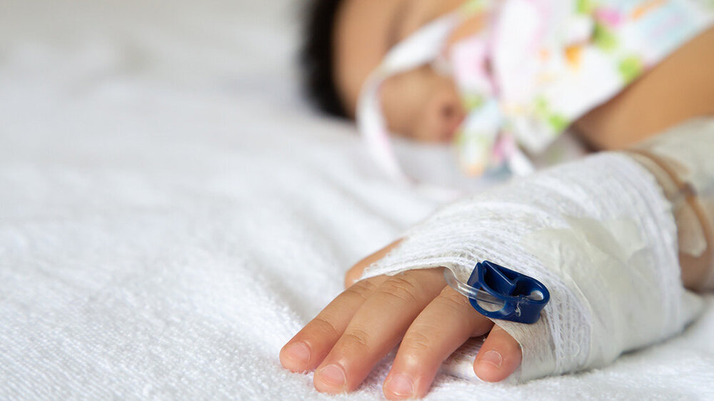 krank_neugeborenes.jpg Hand eines Babys mit einem Verband und einem Zugang liegt auf einem Bett, Gesicht des Babys ist verschwommen im Hintergrund zu sehen