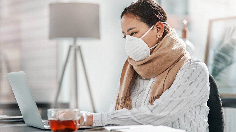 Krank_zur_Arbeit_01.jpg Kranke Frau mit Maske sitzt am PC.