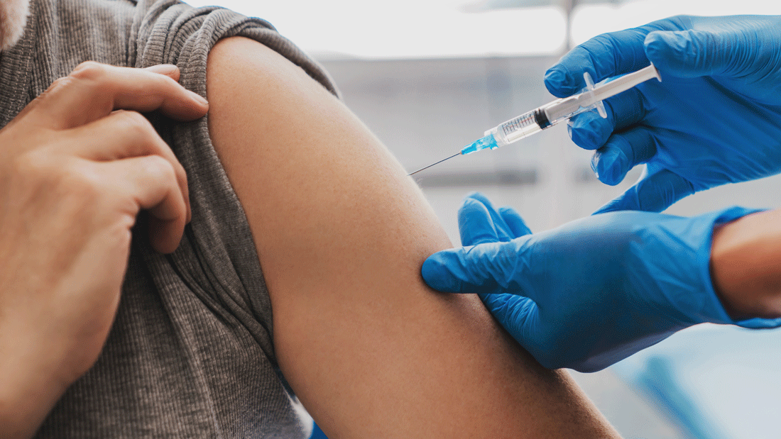 AdobeStock_1004460368.gif Nahaufnahme einer Injektion in den Oberarm: Eine Person zieht den Ärmel hoch, während medizinisches Personal mit blauen Handschuhen eine Spritze ansetzt.