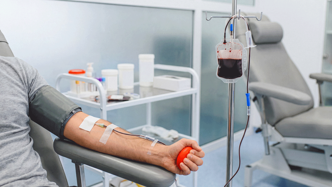AdobeStock_2001481175.gif Eine Person spendet Blut und hält einen roten Ball in der Hand. Über einen Schlauch fließt Blut in einen Beutel am Infusionsständer; die Szene zeigt eine Blutspende in klinischer Umgebung.