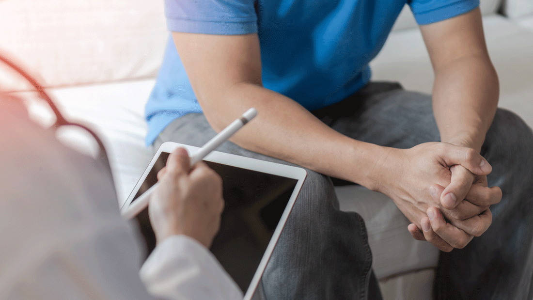 AdobeStock_2184861099.gif Ein Mann im blauen Shirt sitzt mit verschränkten Händen im Gespräch, gegenüber notiert medizinisches Personal etwas auf einem Tablet.