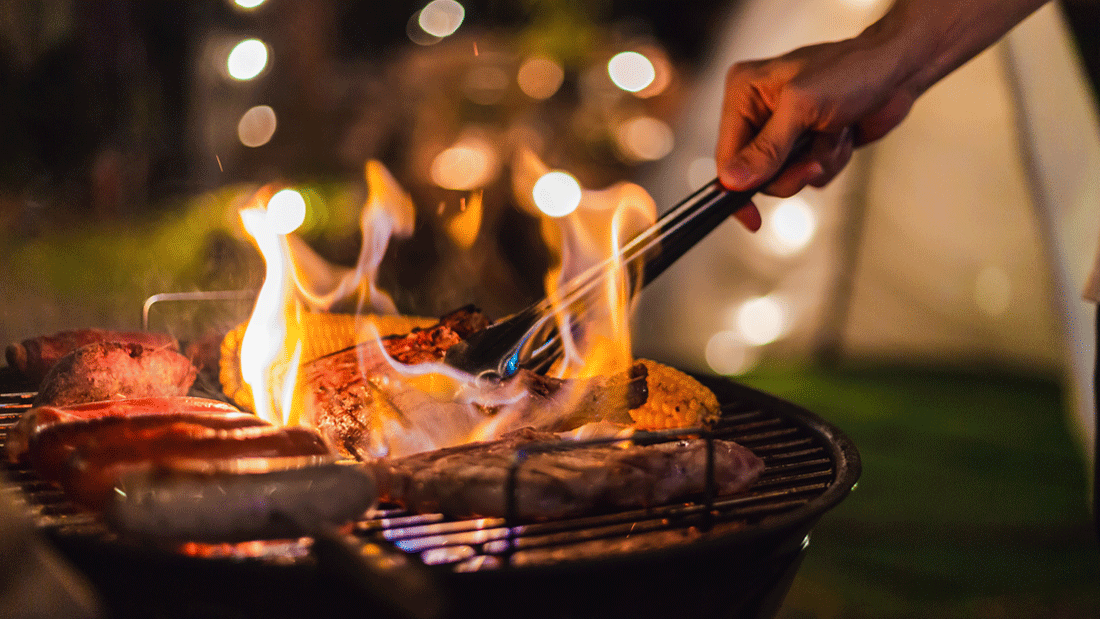 AdobeStock_284530179.gif Nächtliche Grillszene: Eine Hand wendet mit der Zange Fleisch und Mais über offenen Flammen; Würstchen liegen auf dem heißen Rost.