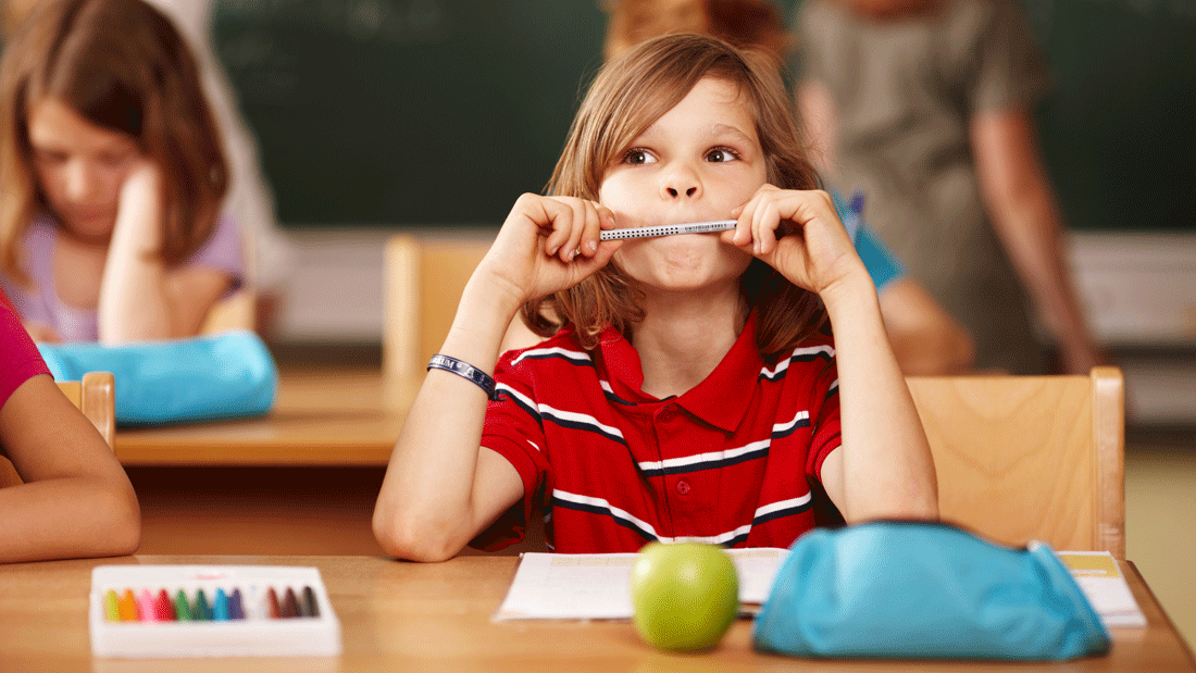 AdobeStock_38834890.gif Dieses Bild zeigt eine Schulszene, in der ein Kind an einem Tisch sitzt, umgeben von Schulmaterialien wie Buntstiften und einer Brotdose. Im Hintergrund sind weitere Kinder und eine Tafel zu sehen.