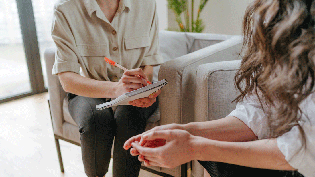 AdobeStock_431034278.gif Das Bild zeigt eine Beratungssituation oder ein Gespräch zwischen zwei Personen. Eine Person hält einen Notizblock und einen Stift, während sie scheinbar Notizen macht. Die andere Person sitzt gegenüber und scheint in das Gespräch vertieft zu sein.