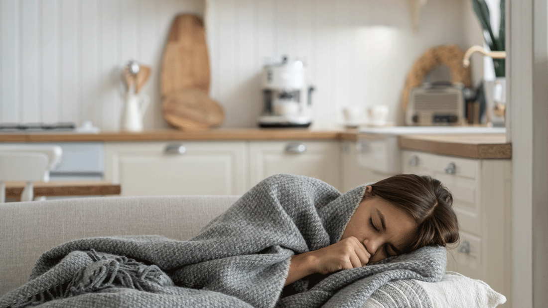 AdobeStock_567629705.gif Das Bild zeigt ein möglicherweise an Grippe erkranktes Kind, das auf einem Sofa liegt und sich in eine warme, graue Decke eingekuschelt hat.