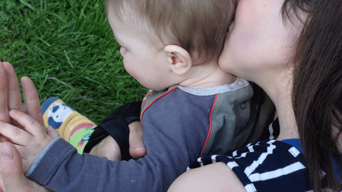 newsimage426674.gif Eine Frau hält ein Baby im Arm und küsst es am Kopf. Beide sitzen im Gras.